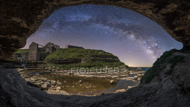 Pedra caverna áspera na praia com pedra ruínas envelhecidas e colinas verdes sob céu noturno com forma leitosa e estrelas — Fotografia de Stock