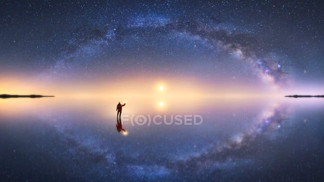 Silhueta de homem anônimo em pé na superfície de reflexão da água e estendendo a mão para o céu estrelado colorido noite com forma leitosa — Fotografia de Stock
