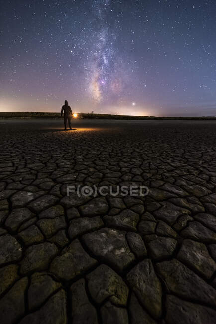 Back view of anonymous man standing on empty road with lantern on a colorful nigh sky with milky way on background — Stock Photo