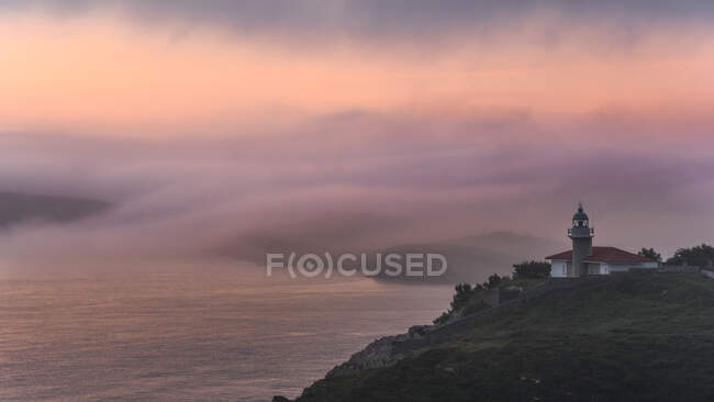 Маленькая старая церковь на зеленом одиноком холме на берегу океана во время туманного утра с облачным красочным небом на заднем плане — стоковое фото