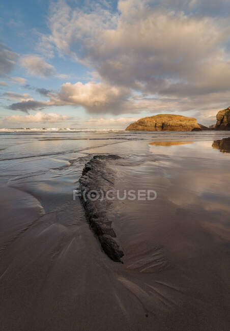Big rocky formations on sandy empty beach of ocean with cloudy bright evening sky on background — Stock Photo