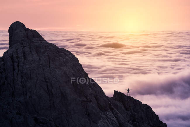 Silhouette of unrecognizable person stretching arms standing on peak of rough cliff with colorful clouds on background during sunset — Stock Photo