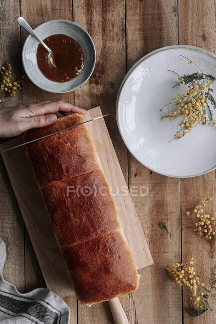 From above top view cropped unrecognizable person hand with rectangle loaf of fresh Brioche bread on wooden table with bowl of jam and knife — Stock Photo