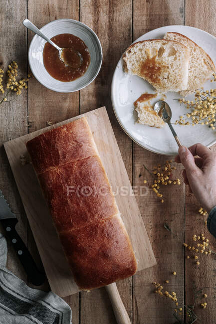 From above top view cropped unrecognizable person hand with rectangle loaf of fresh Brioche bread on wooden table with bowl of jam and knife — Stock Photo