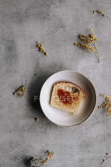 Вид зверху на свіжий шматочок хліба Бріоша, намазаного коричневим варенням на тарілці на сірому столі з квітами — стокове фото