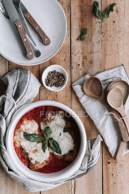 From above ceramic bowl with pieces of roasted chicken with Parmesan and basil in tomato sauce placed on timber table near napkin and utensils — Stock Photo