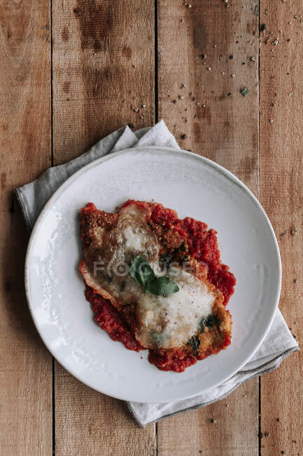 Top view of piece of roasted chicken with Parmesan and leaf of basil placed on tomato sauce on plate over napkin and wooden table — Stock Photo