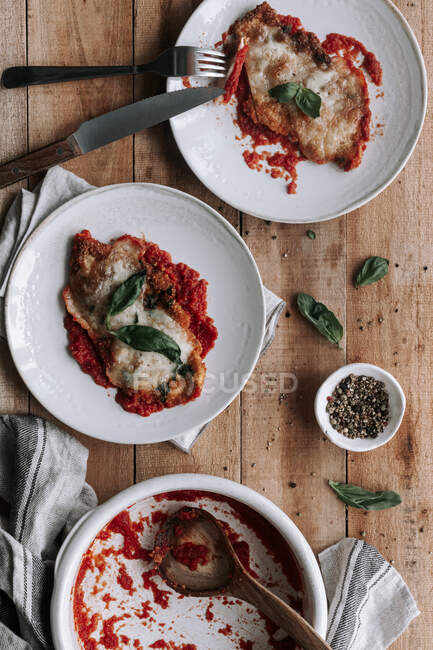 Top view of bowl and plate with yummy chicken with cheese and basil roasted in tomato sauce placed near napkin and spices on wooden table — Stock Photo