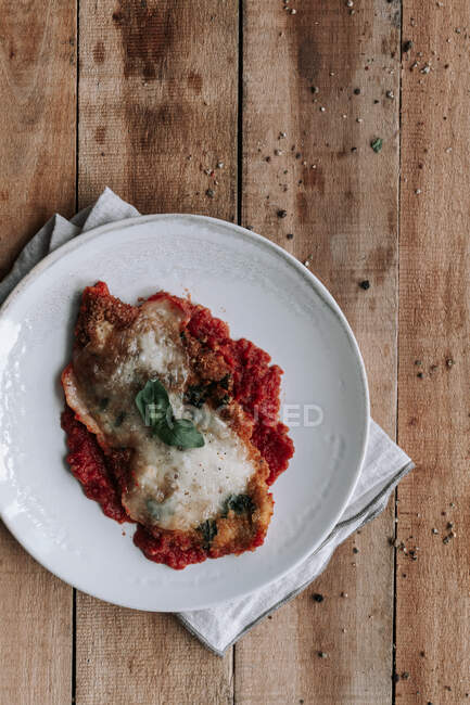 Top view of piece of roasted chicken with Parmesan and leaf of basil placed on tomato sauce on plate over napkin and wooden table — Stock Photo