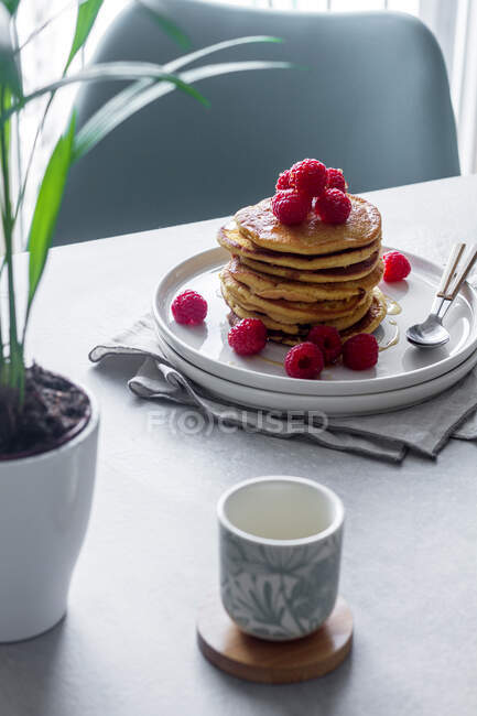De cima prato de deliciosas panquecas com framboesas colocadas em guardanapo perto de copo vazio e vaso de planta na parte da manhã — Fotografia de Stock
