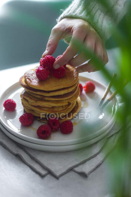 Persona irriconoscibile in maglia maglione mettendo lamponi freschi sulla cima di gustosa pila frittelle durante la colazione a casa — Foto stock