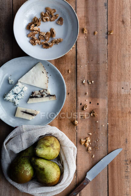 De cima saco de algodão com peras frescas e prato com nozes colocadas perto de queijo e faca na mesa de madeira — Fotografia de Stock