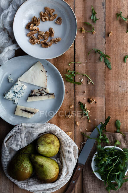 Top view of plates with walnuts and cheese placed near cotton bag with ripe pears and bowl with fresh arugula on lumber table during salad preparation — Stock Photo