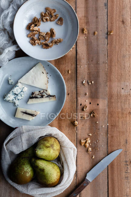 Сверху хлопчатобумажный мешок со свежими грушами и тарелкой с грецкими орехами помещен рядом с сыром и ножом на деревянном столе — стоковое фото