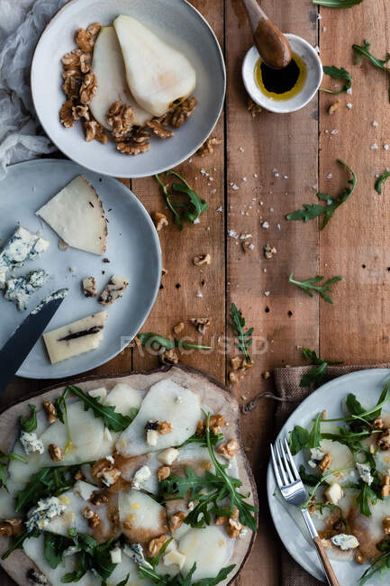 De pratos acima de pêra deliciosa e salada de nozes com queijo e arugula colocados na mesa de madeira perto de ingredientes de cozinha — Fotografia de Stock