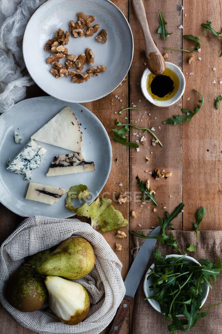 Top view of plates with walnuts and cheese placed near cotton bag with ripe pears and bowl with fresh arugula on lumber table during salad preparation — Stock Photo