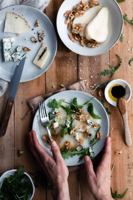 Вид зверху анонімної людини, що кладе тарілку грушевого салату з руколою на дерев'яний стіл біля сиру та волоських горіхів на кухні — стокове фото