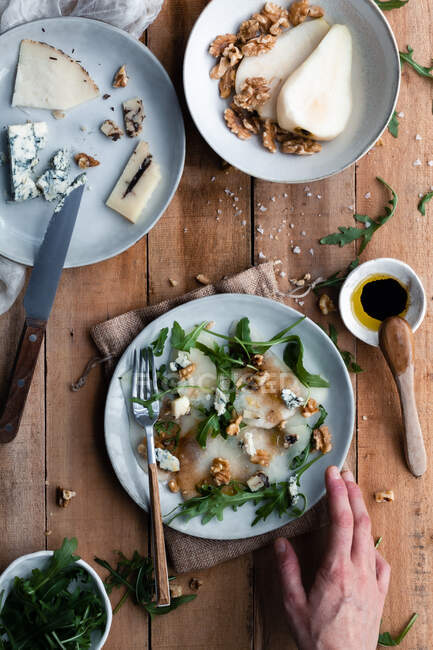 Top view of anonymous person putting plate of pear salad with arugula on timber table near cheese and walnuts in kitchen — Stock Photo