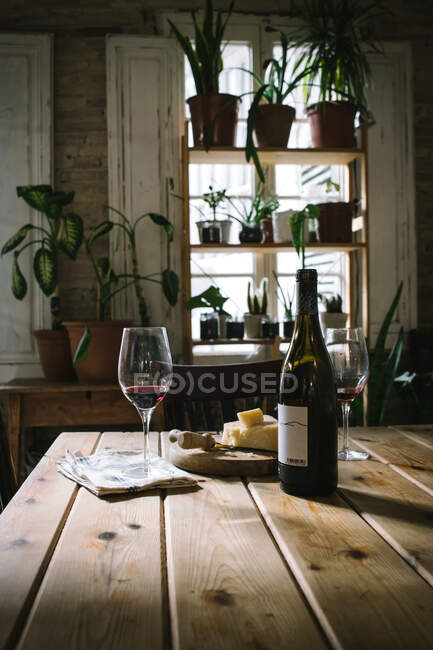 Open bottle and glasses with red wine placed near cheese on wooden table in rustic restaurant with potted green plants on window — Stock Photo
