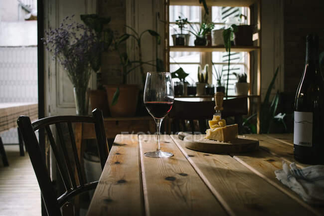 Open bottle and glasses with red wine placed near cheese on wooden table in rustic restaurant with potted green plants on window — Stock Photo