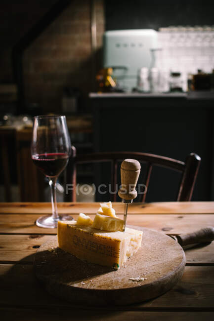 Pedazo de delicioso queso con cuchillo servido en tablero de madera colocado cerca de un vaso de vino tinto en una mesa de madera en un bar rústico - foto de stock