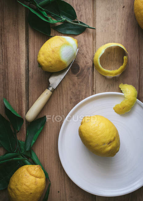 Вид сверху очищенных и свежих лимонов на тарелках на деревянном столе с зелеными листьями — стоковое фото