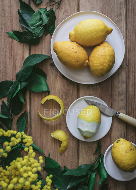 Vue du dessus des citrons pelés et frais sur des assiettes sur une table en bois avec des feuilles vertes et des fleurs jaunes — Photo de stock