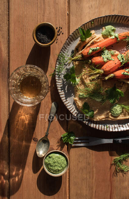 Vista superior de fatias de pão fresco espalhadas com hummus na placa com cenouras laranja assadas decoradas com molho verde na mesa de madeira — Fotografia de Stock