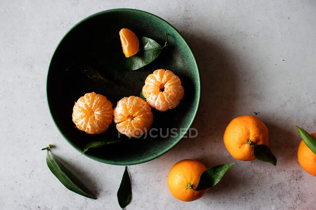 Vista superior del tazón de cerámica verde con mandarinas peladas frescas colocadas sobre una mesa blanca cerca de frutas sin pelar con hojas verdes - foto de stock