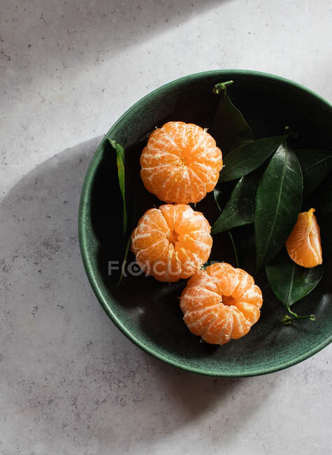 Vista dall'alto di ciotola di ceramica verde con mandarini freschi pelati disposti su tavolo bianco vicino a frutti non pelati con foglie verdi — Foto stock
