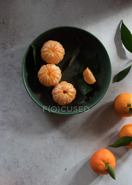Vista superior del tazón de cerámica verde con mandarinas peladas frescas colocadas sobre una mesa blanca cerca de frutas sin pelar con hojas verdes - foto de stock
