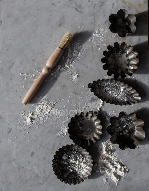 Top view of various metal baking molds and small kitchen brush on marble table sprinkled with white flour — Stock Photo