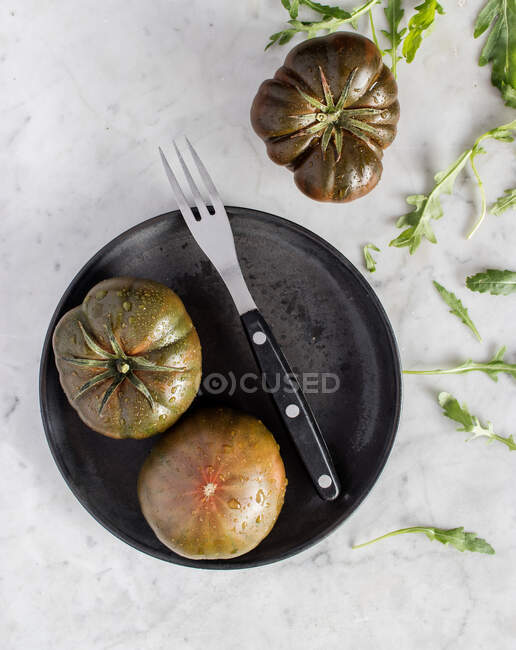 Вид сверху больших незрелых зеленых помидоров на черной тарелке с металлической вилкой и кусочком сыра на тарелке — стоковое фото
