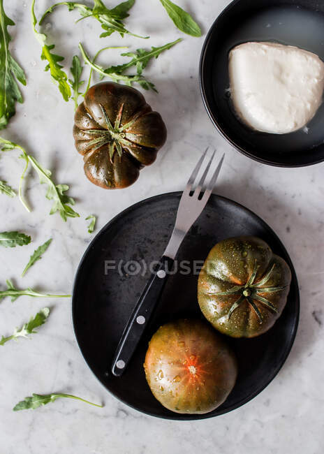 Вид сверху больших незрелых зеленых помидоров на черной тарелке с металлической вилкой и кусочком сыра на тарелке — стоковое фото