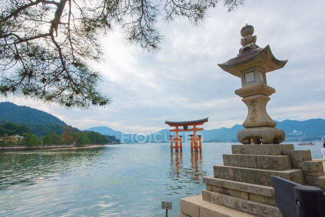 Peaceful Japanese landscape with built over water gate and stone religious sculpture against cloudy sky framed with branches of pine tree — Stock Photo