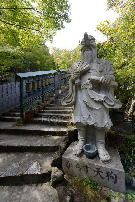 Старая каменная буддийская статуя рядом с выветренной каменной лестницей в парке с зелеными деревьями в солнечный летний день в Японии — стоковое фото