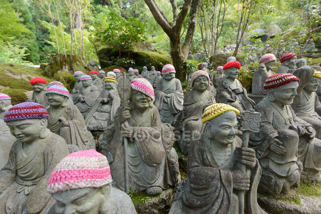 Многие каменные буддийские скульптуры, одетые в красочные вязаные шляпы в зеленом лесу в Японии — стоковое фото