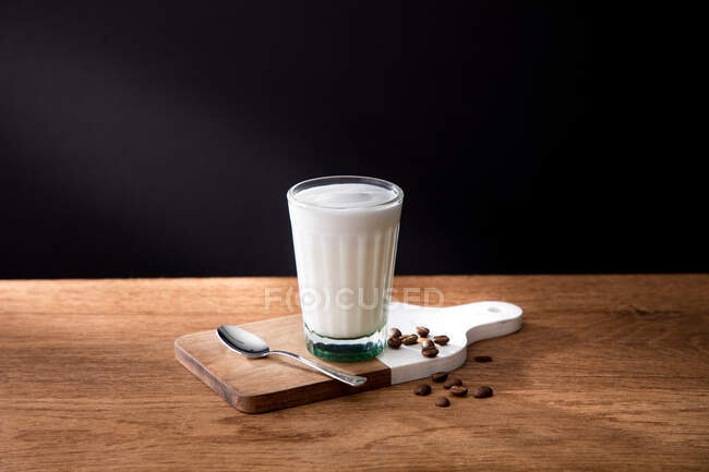 Copo de leite fresco colocado em tábua de madeira com colher e grãos de café em mesa de madeira com fundo preto — Fotografia de Stock