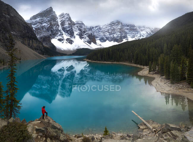 From above side view of unrecognizable male traveler standing on shore of beautiful lake with clear turquoise water surrounded by snow covered rocky mountains and coniferous forest reflected in water in cloudy day in Canada — Stock Photo