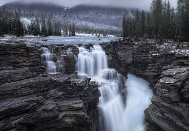 Espectacular paisaje con hermosa cascada en cascada entre montañas rocosas cubiertas de bosque de coníferas y nieve en Canadá - foto de stock