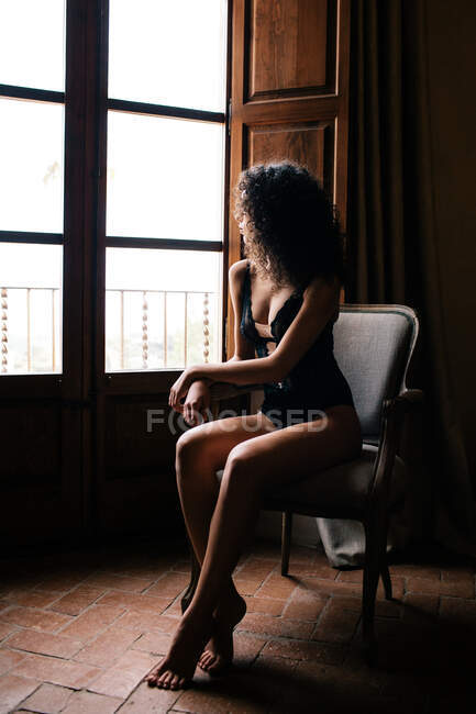 Vista laterale del corpo pieno sensuale giovane donna in biancheria intima seduta su comoda sedia e guardando fuori finestra in camera vintage scuro — Foto stock