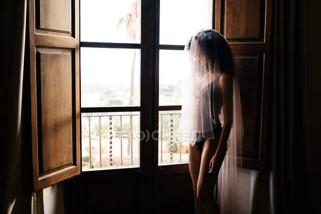 Mujer delgada irreconocible en ropa interior y velo translúcido de pie cerca de la ventana con persianas abiertas en habitación retro oscura - foto de stock