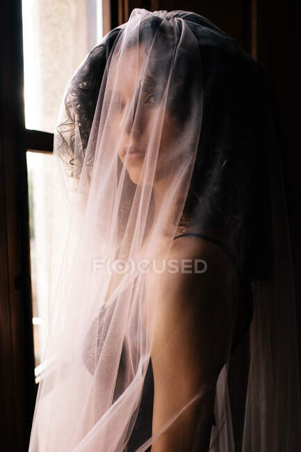 Mujer mirando a la cámara con velo translúcido de pie cerca de la ventana con persianas abiertas en habitación retro oscura - foto de stock