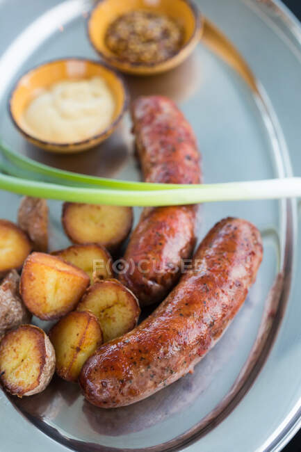 Dall'alto pezzi di deliziose patate alla griglia e succose salsicce con scalogno fresco poste su piastra metallica vicino a ciotole con salse in mensa — Foto stock