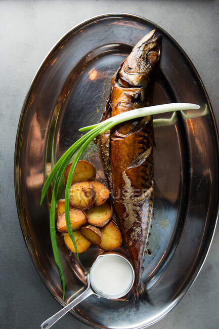 Vista superior de delicioso pescado asado y papas servidas con cebolleta madura y salsa de crema en el plato de metal en el restaurante - foto de stock