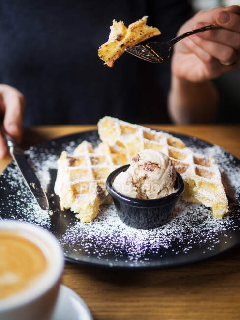 Unrecognizable person using form and knife to cut sweet waffles near bowl of ice cream and cup of coffee on table in cafe — Stock Photo