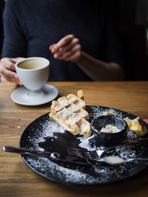 Тарелка с остатками вафель и мороженого на столе в столовой рядом с неузнаваемым человеком, пьющим кофе — стоковое фото