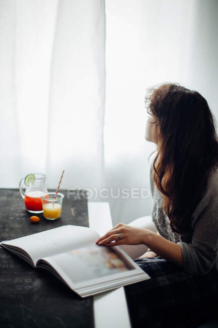 Vue latérale d'une jeune femme en tenue décontractée lisant un livre assis à table avec du jus de fruits dans une pièce lumineuse — Photo de stock