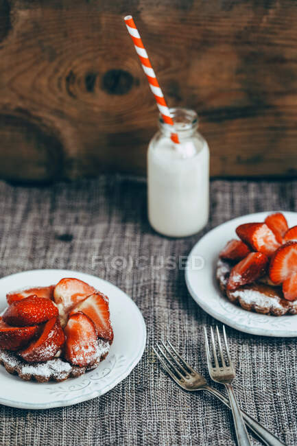 From above of delicious homemade waffles with ripe strawberry and sugar powder served on white plates on table with milk drink in glass bottle — Stock Photo