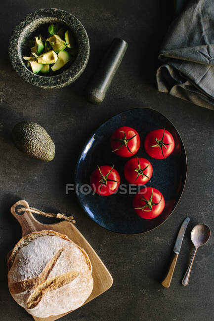 Top view of mortar with chopped avocado and plate with tomatoes placed near various utensils and fresh bread on gray table — Stock Photo